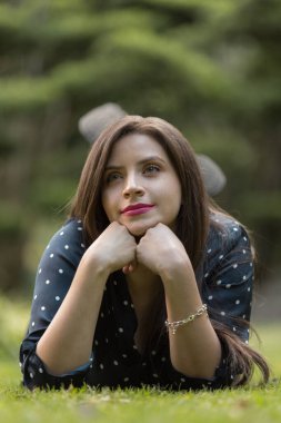 Uzun saçlı ve makyajlı genç beyaz tenli kadın, elleri çenesinde çimenlerde uzanıyor, gülümsüyor ve bilezik takıyor, üzerinde beyaz noktalı siyah bir bluz var.