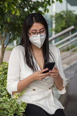 Uzun siyah saçlı, gözlüklü ve maskeli Latin bir kadın cep telefonunu kullanarak bitkilerin yanına oturur.