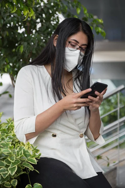 Duvarda bitki ve yapraklarla otururken cep telefonu kullanan bir kadın. Maske ve gözlüklerle virüse karşı koruma