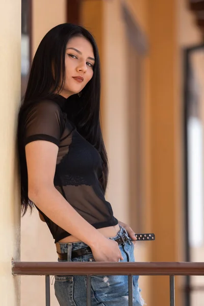 美丽的年轻拉丁女子 一头直发 靠着墙 穿着一件透明的衬衫 牛仔裤和一条黑带 — 图库照片