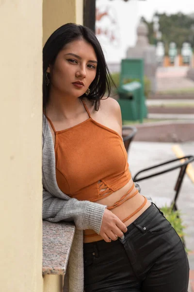 年轻的拉丁女人 黑头发 靠着墙站着 身穿橙色上衣 黑色裤子和灰色夹克 露出腰部 阳光灿烂 — 图库照片
