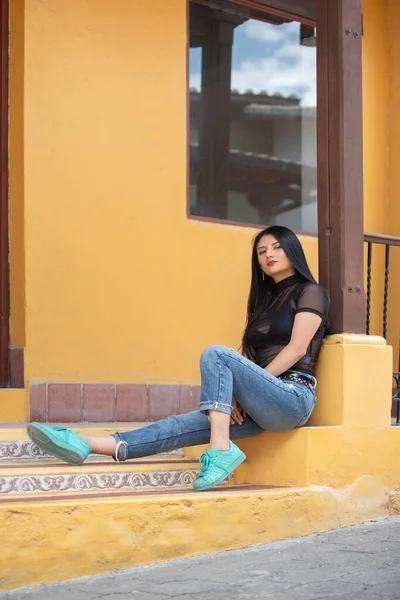 Young Latina woman with long black hair, sitting on a step, wearing a transparent blouse, jeans and turquoise shoes, home interior