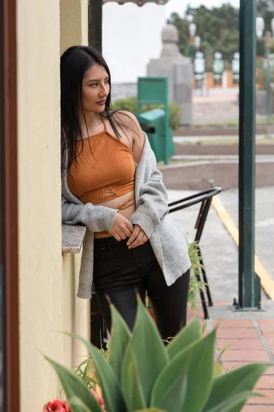 Young Latina woman with long black hair, standing leaning against the wall, wearing an orange top, black pants and gray jacket, exterior