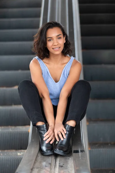 beautiful young brunette latina woman with short hair and curls, happily sitting on a handrail, wearing blue blouse, black pants and boots