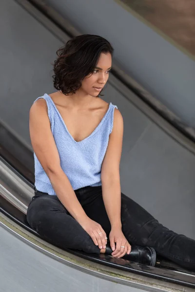 young brunette latina woman with short hair with curls, sitting and distracted on a handrail of electric bleachers, wearing blue blouse, black pants and boots