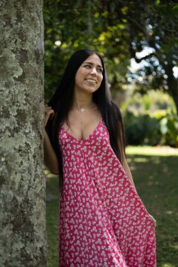 Uzun kırmızı çiçekli elbiseli güzel bir kadın güneşli bir bahçede ağaca yaslanırken sıcak bir şekilde gülümsüyor. Uzun siyah saçları doğal akarken çevreci resmiyle çevrili bu açık havada neşe, zarafet ve cazibesini sunuyor..