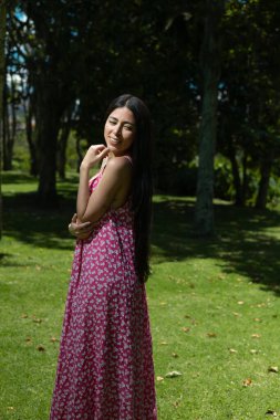 Akan kırmızı elbiseli güzel bir kadın zarif bir şekilde bir ağaca yaslanıyor, gözleri kapalı, yemyeşil bir bahçedeki güneşin sıcaklığının tadını çıkarıyor..