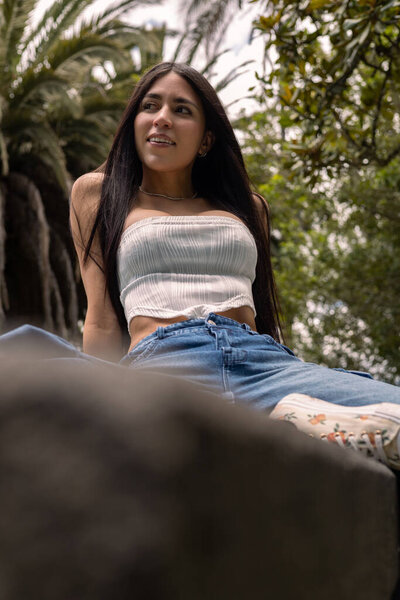 Красивая молодая женщина с длинными темными волосами изящно сидит на деревенской скамейке в освещенном солнцем саду. Нося джинсы в джинсах и белый топ, она излучает очарование, элегантность и расслабленную уверенность, наслаждаясь мирной атмосферой сельской местности.