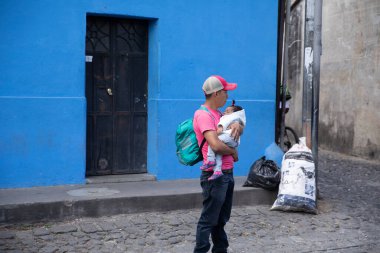 Mavi şapkalı ve beyaz tişörtlü, sokakta sırt çantalı bir çocuk.