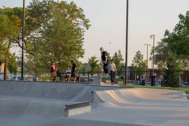 Kaykaycılar ve scooterlar numaralarını güneşli bir yaz gününde halka açık olan açık bir kaykay parkında yapıyorlar.