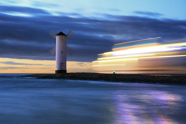 Luxury boat in motion at Baltic sea. Cold icy evening. Night sea scene near frozen beach during suset. Beautiful sunset over a windmill-shaped lighthouse. Swinoujscie, Poland.