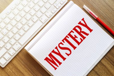MYSTERY yazılı beyaz bir defter kırmızı işaretli beyaz bir klavyenin yanındaki ahşap bir yüzeye uzanıyor.