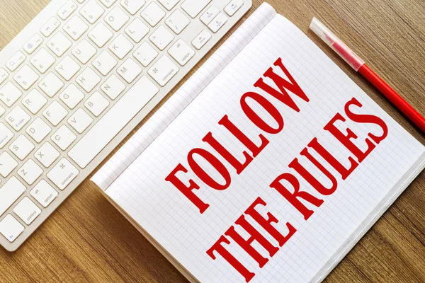 a white notebook with the text Follow The Rules lies on a wooden surface near a white keyboard with a red marker