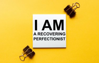 Kırtasiyeli sarı bir arka planda iyileşmekte olan bir mükemmeliyetçiyim yazan beyaz bir kağıt.