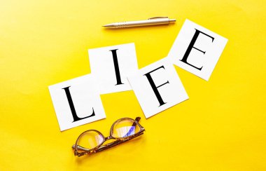 Yazılı beyaz kağıt. Sarı arka planda gözlük ve kalemle yaşam.