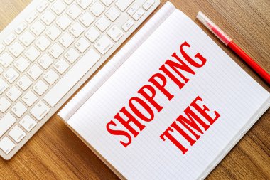 Metin Alışveriş Zamanı yazılı beyaz bir defter kırmızı işaretli beyaz bir klavyenin yanındaki ahşap bir yüzeye uzanır.