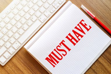 Metni olan beyaz bir defter kırmızı işaretli beyaz bir klavyenin yanındaki ahşap bir yüzeyde olmalı.