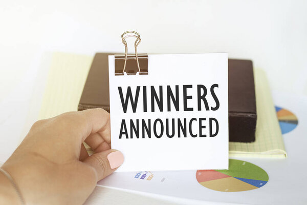 Рука женщины держит лист бумаги с текстом Победители объявлены на фоне графиков и блокнота