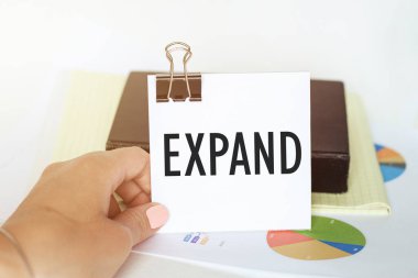 Kadın eli, arka planda grafik ve not defterinin üzerinde ExPAND yazılı bir kağıt parçası tutuyor.