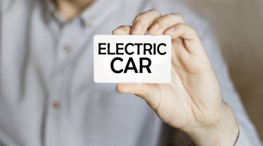 Mavi gömlekli bir işadamı elinde elektrikli araba yazan bir kart tutuyor.