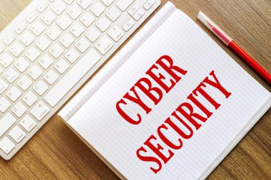 Beyaz bir defterde Siber Güvenlik yazılıydı. Beyaz bir klavyenin yanında, üzerinde kırmızı işaret olan tahta bir yüzey vardı.
