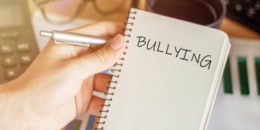 Erkek elinde bir defter tutuyor. Ofis masasının arka planında zorbalık yazılı bir defter.