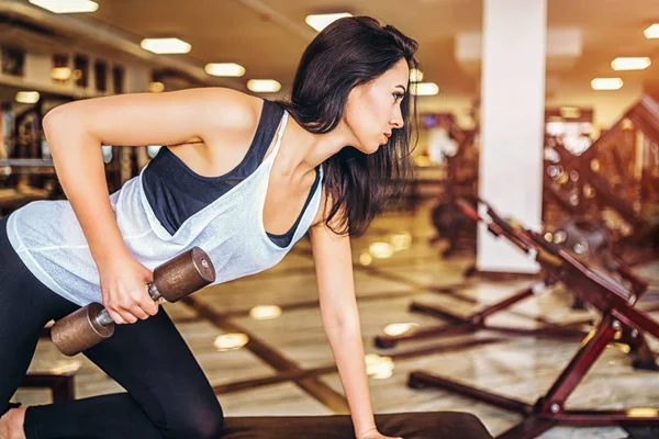 Woman leaning on bench while training with dumbbell in gym