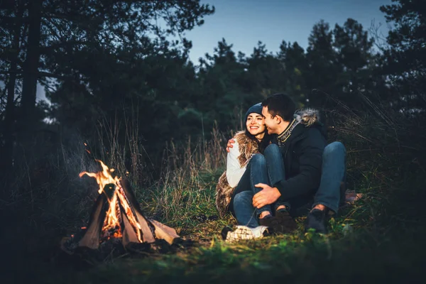 Pretty young couple relaxing near bonfire in the forest at evening time