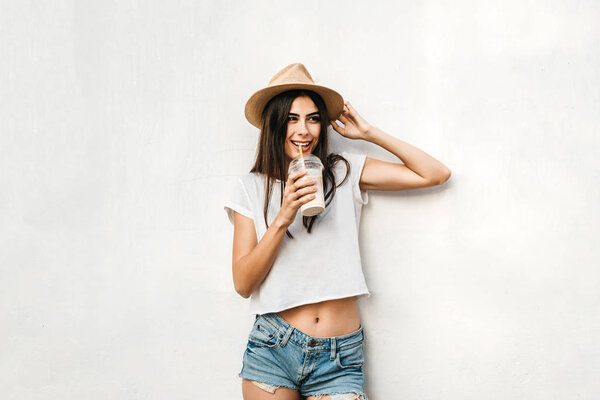 Pretty brunette girl in hat with coffee and white wall behind, copy space