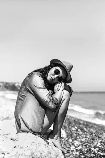 Menina morena bonita em chapéu sentado na pedra perto do mar . — Fotografia de Stock