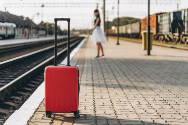 Beyaz etekli, kırmızı valizli, trenini bekleyen ve tren istasyonunda yürüyen esmer bir kadın yolcu.