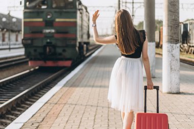 Beyaz etekli, kırmızı valizli, trenini bekleyen ve tren istasyonundaki trene el sallayan esmer bir kadın yolcu.