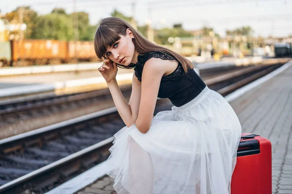 身穿白色短裙 头戴红色行李箱的女性黑发旅行者在候车站等火车 — 图库照片