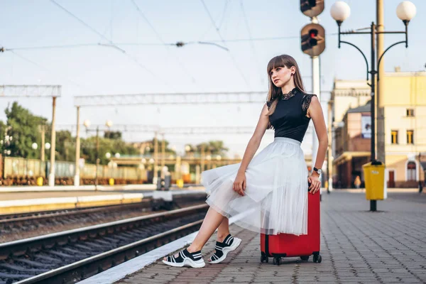 女性ブルネット旅行者とともに赤スーツケースでホワイトスカート待っていますのために列車上の鉄道駅 — ストック写真