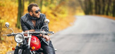 Güneş gözlüklü, deri ceketli, sakallı, vahşi bir adam ormanda bir motosikletin üzerinde oturuyor..