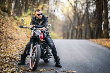 Güneş gözlüklü, deri ceketli, sakallı, vahşi bir adam ormanda bir motosikletin üzerinde oturuyor..