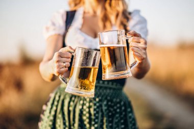 Kızların elleri dirndl 'de, geleneksel festival elbisesi içinde, kocaman köpüklü bir bardak bira doldururken, arka planda bulanıklık var. Ekim Festivali, Aziz Patrick Günü, uluslararası bira günü konsepti