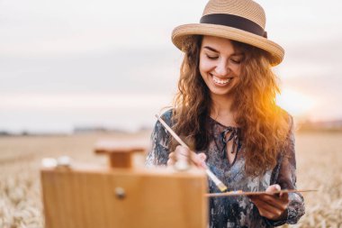 Güzel kız ressam buğday tarlasında resim yapıyor. Kadın gülümsüyor, elinde bir fırça ve palet tutuyor, önünde bir sehpa duruyor. Arkasında turuncu güneş ışığı var..