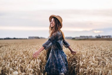 Kıvırcık saçlı güzel bir genç kadının yakın plan portresi. Buğday tarlasında duran şapkalı ve elbiseli kadın..