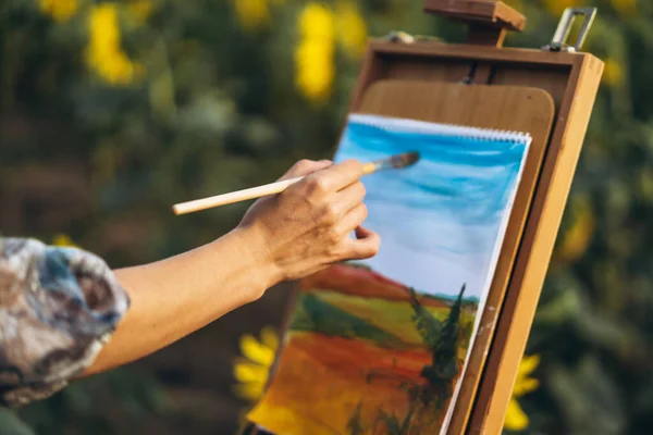 Close up hands of female artist holding brush and draws a picture of the field. Blurred background with easel in sunflower field.