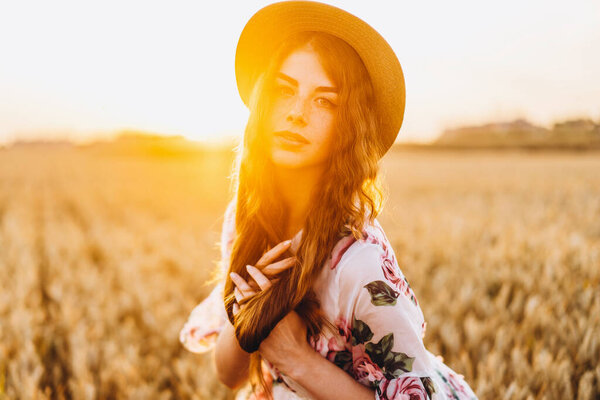 Portrait of a beautiful young woman with curly hair and freckles face. Woman in dress and hat posing in wheat field at sunset and looking at camera.