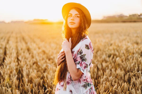 一个有着卷发和雀斑的漂亮年轻女子的画像 日落时分 身穿连衣裙 头戴帽子的女人站在麦田里 看着镜头 — 图库照片
