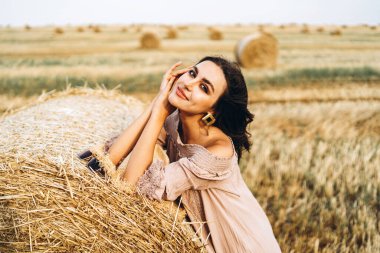 Gözleri kapalı, gülümseyen güzel bir kadının yakın plan portresi. Esmer, bir saman balyasına yaslandı. Arka planda bir buğday tarlası.