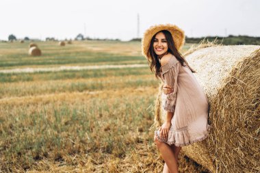 Güneş gözlüklü ve saman balyalarının yanındaki buğday tarlasında poz veren uzun saçlı güzel bir genç kız. Yazlık elbiseler içinde mutlu esmer.