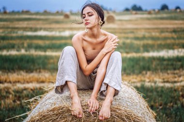 Ilık bir sonbahar gününde saman balyalarında oturan keten pantolonlu ve çıplak omuzlu bir esmer. Kameraya bakan kadın. Arkasında bir buğday tarlası var..