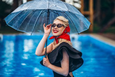 Güneş gözlüklü, kırmızı dudaklı, siyah, klasik mayo ve kırmızı eşarplı, şemsiyeli ve yüzme havuzunda iyi eğlenceler.