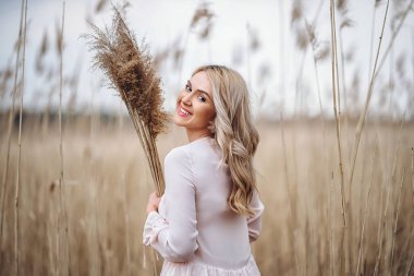 Uzun sarı kıvırcık saçlı, güzel bir kızın fotoğrafı. Sazlık tarlasında duruyor ve elinde yüksek kamış dalları var.
