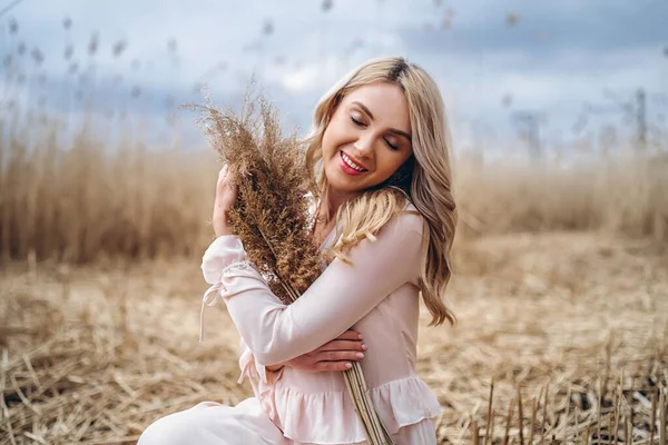 照片上是一个漂亮的微笑着的女孩 一头长长的金发卷曲在淡淡的长长的芦苇里 手里拿着高高的芦苇枝条 — 图库照片