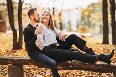 Ormandaki ahşap bankta oturan sevgi dolu genç bir çift. Erkek ve kadın, sonbahar ağaçlarının arka planında sarılıp gülümsüyor..