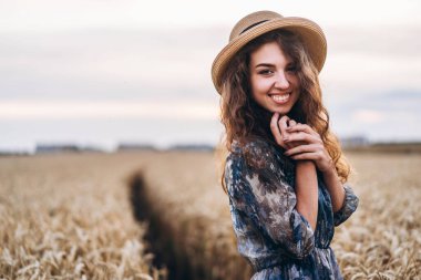 Kıvırcık saçlı güzel bir genç kadının yakın plan portresi. Buğday tarlasında duran şapkalı ve elbiseli kadın..